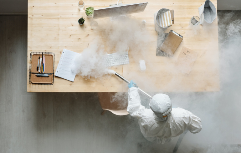 Professionelle Geruchsbeseitigung in Reutlingen. Eine Person im weißen Schutzanzug führt eine professionelle Geruchsneutralisierung mit dichtem Nebel an einem Schreibtisch durch.